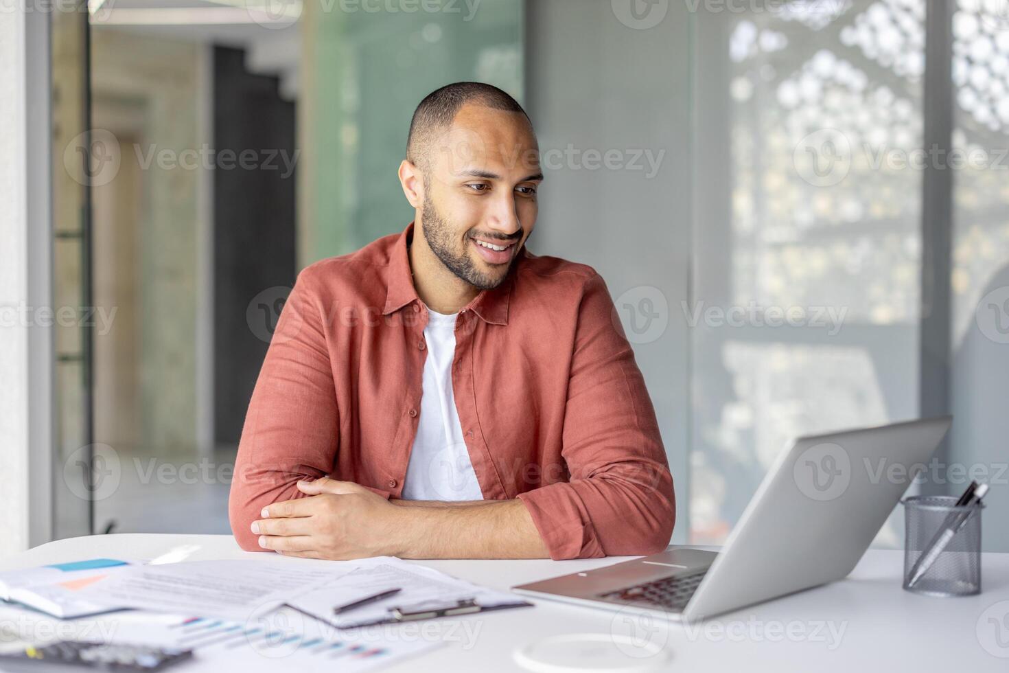 Young adult man sitting at a desk in a modern office is having a call on his laptop, reviewing documents and smiling during the conversation photo