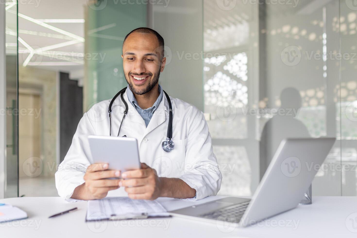 Successful experienced doctor sitting at desk inside the clinic office and using a tablet computer. Man in a white medical coat, using an application for online communication and consulting patients. photo