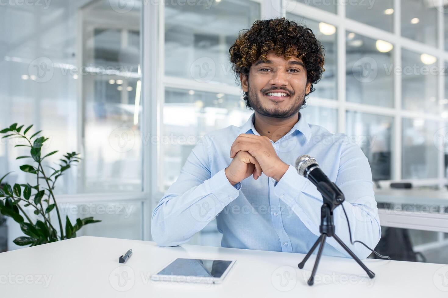 Confident businessman sitting at an office desk with a microphone, ready for a podcast interview. Office setting with modern decor in the background. photo