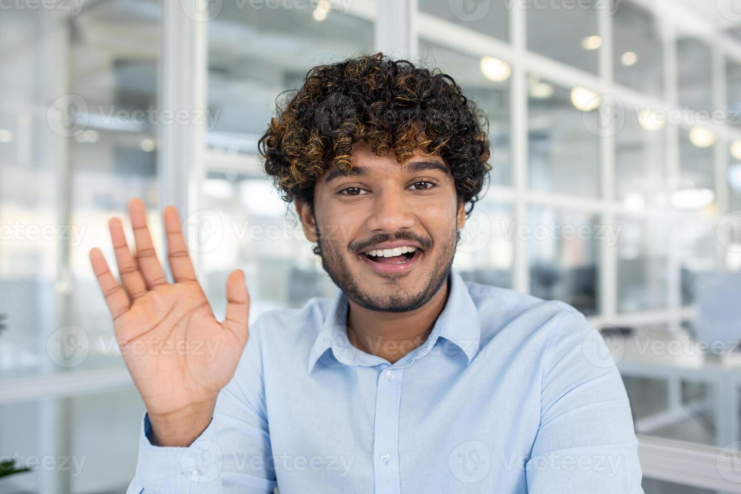 Young, curly-haired man in a light blue shirt warmly greeting with a wave, exuding friendliness and positivity in a bright, contemporary office environment. photo