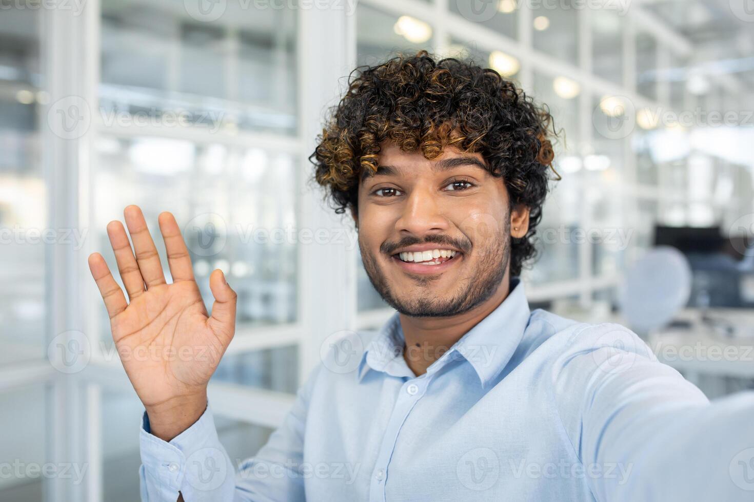 A cheerful young man with curly hair greeting enthusiastically in a modern office environment. He's wearing a light blue shirt and smiling at the camera. photo