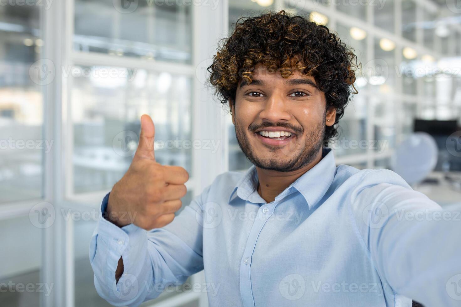 Confident young man with curly hair showing a thumbs up while taking a selfie at a bright, well-lit office setting. Positive and professional vibe. photo
