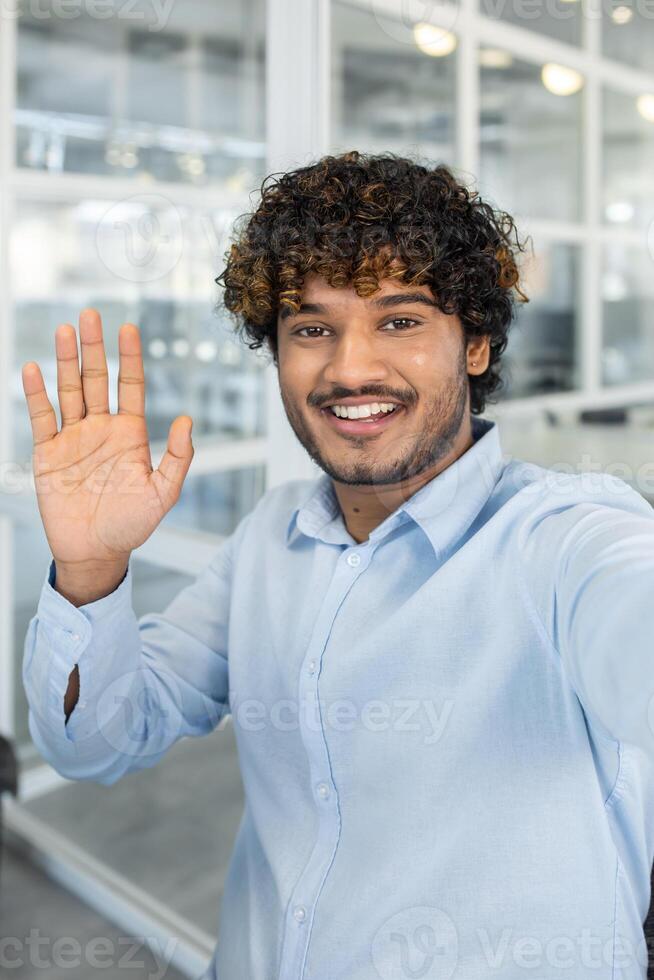 Cheerful young adult male with curly hair waving at the camera, possibly during a call, in a brightly lit modern office setting. photo