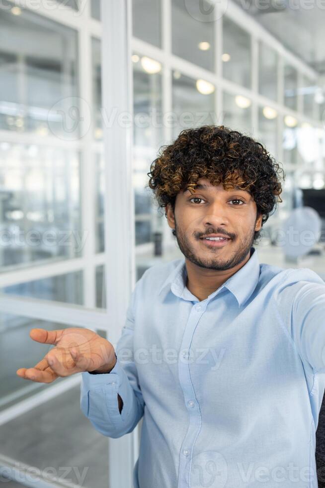 A smiling young man with curly hair makes a call from a modern office. He's looking at the camera, engaging in a friendly conversation. photo