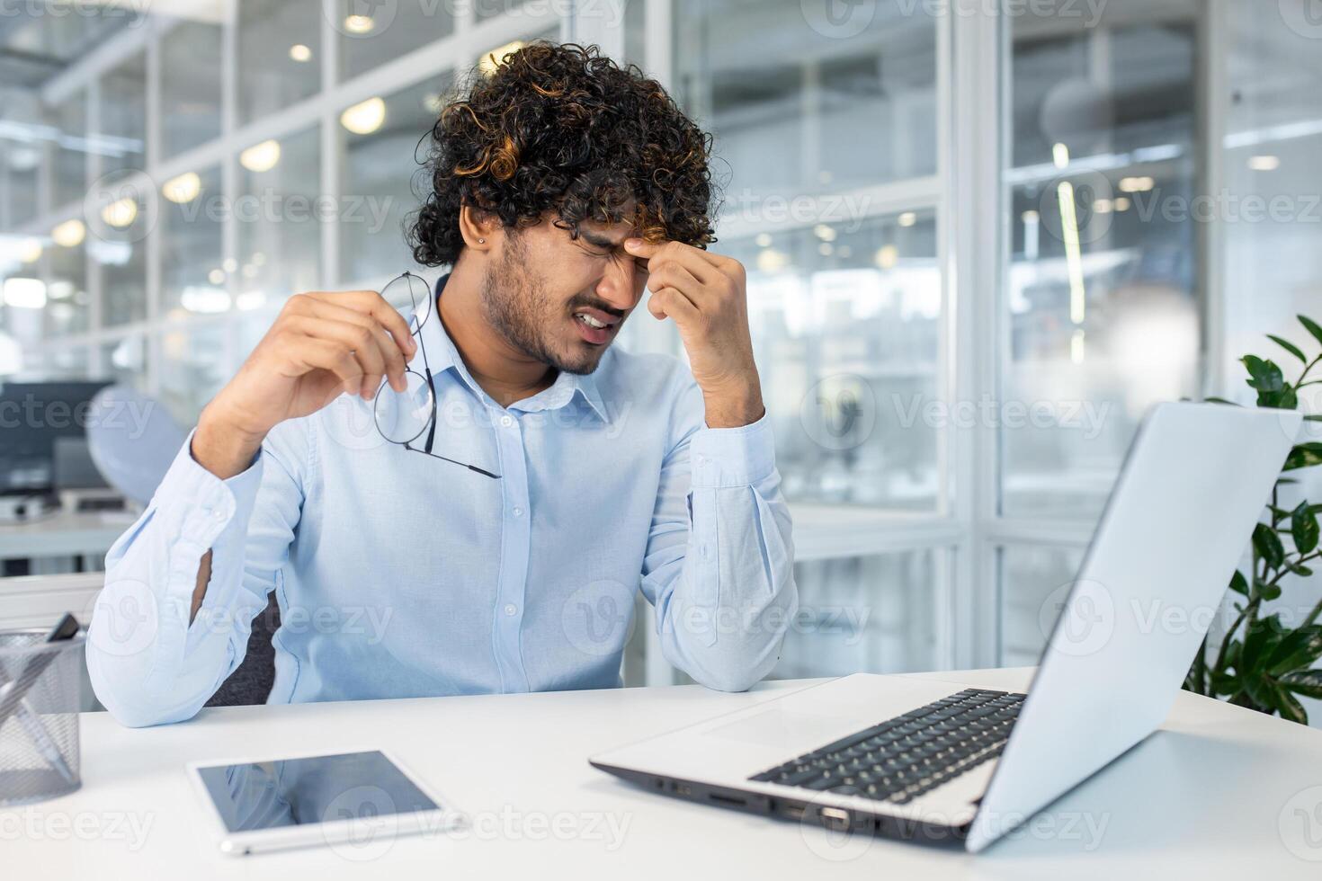 A young man sits at his office desk, visibly frustrated, rubbing his temples in an attempt to alleviate stress or a headache. An open laptop and digital tablet are on the desk. photo