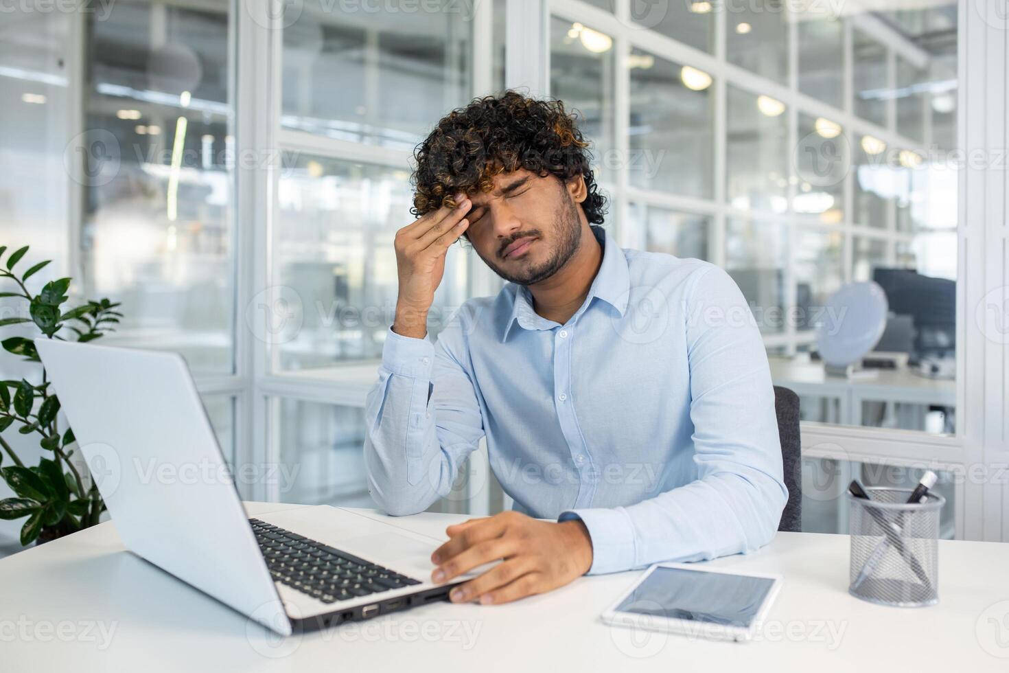 A young man in a blue shirt feels a headache while working on his laptop in a modern office setting, signalling stress and fatigue. photo