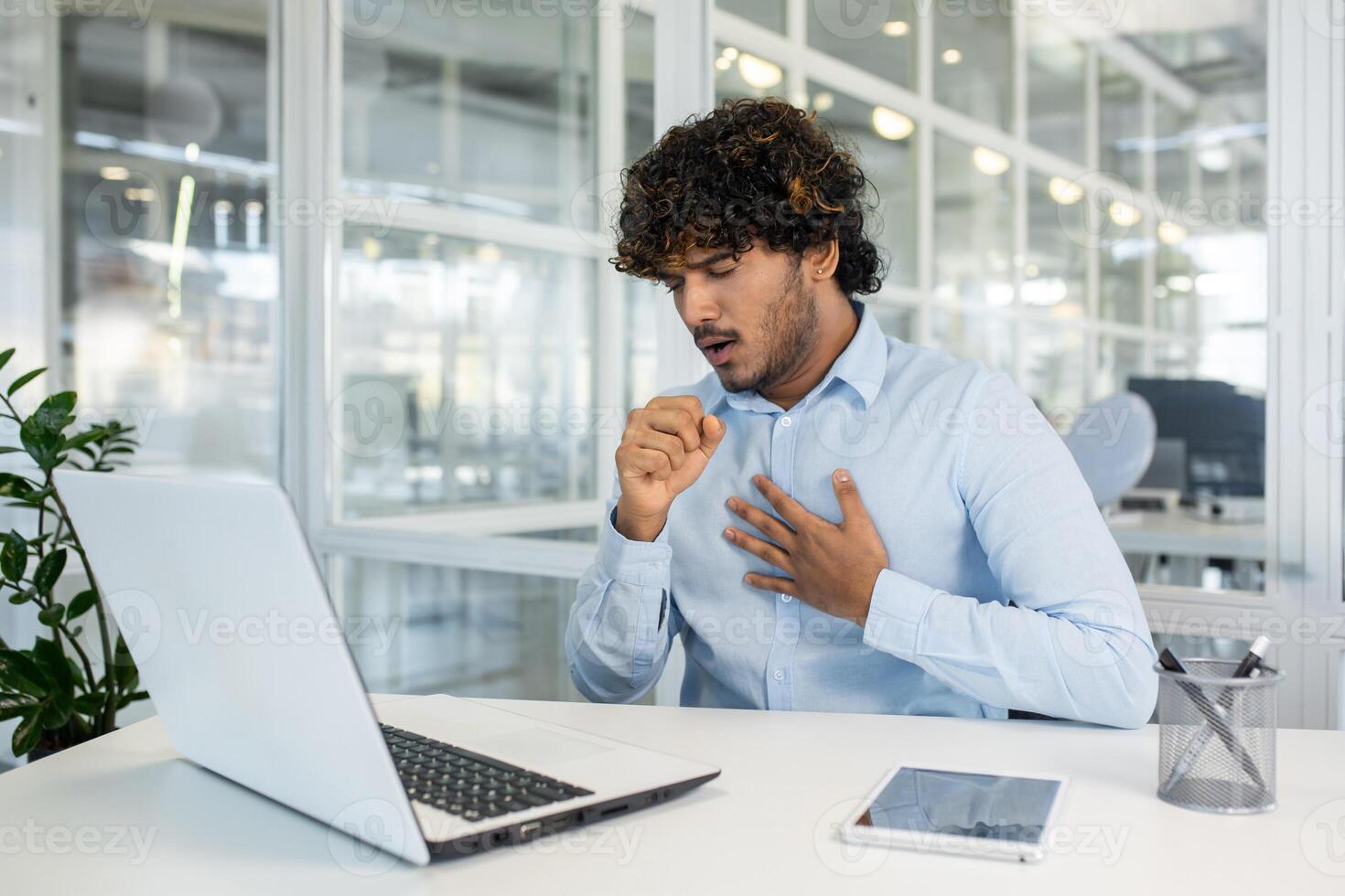 A young man in a light blue shirt feels a sudden cough while working on his laptop in a modern office setting. Concern and discomfort are evident as he addresses his health at work. photo