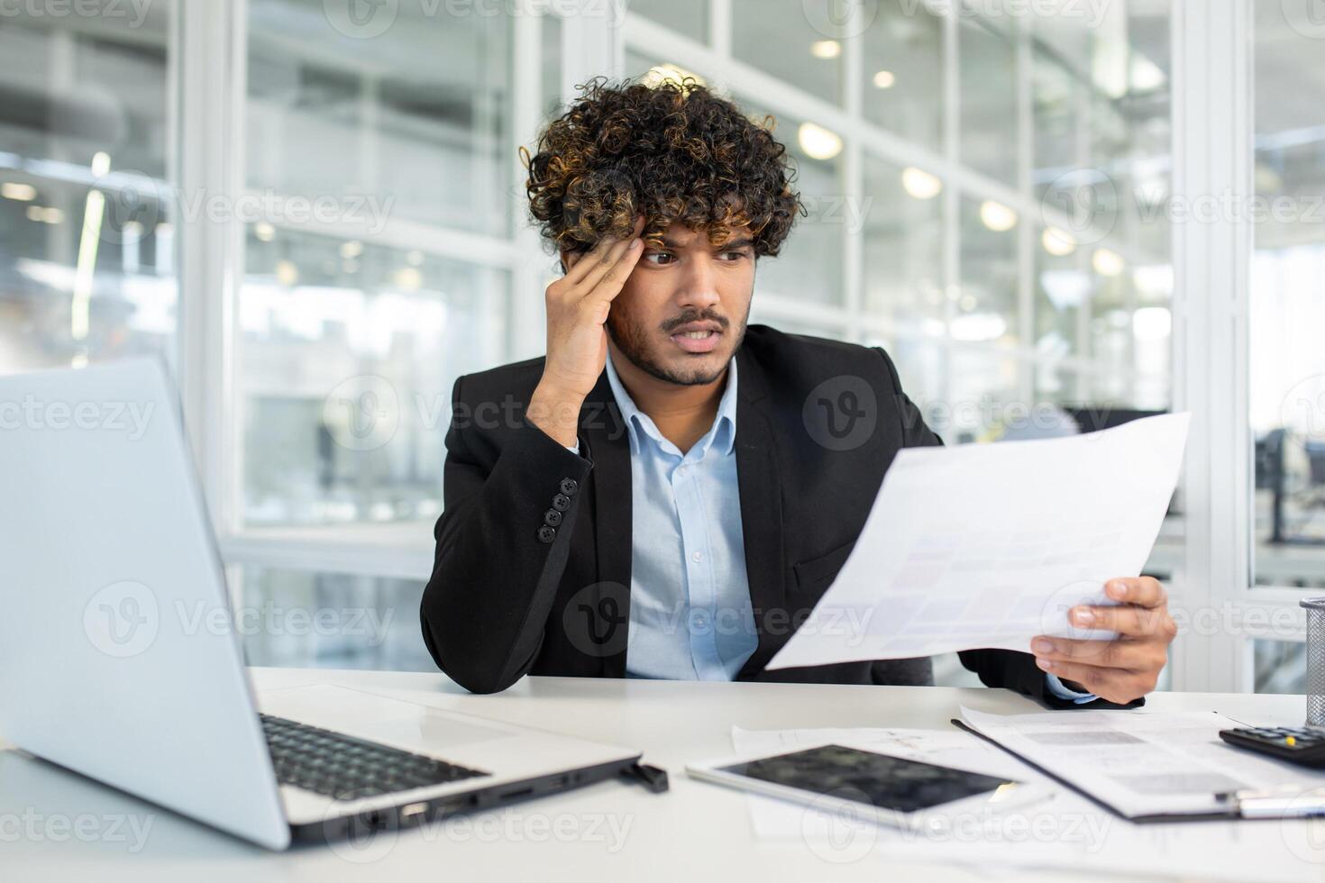 A stressed business professional in suit looks over important documents at his workspace in a bright, modern office setting, surrounded by technology. photo