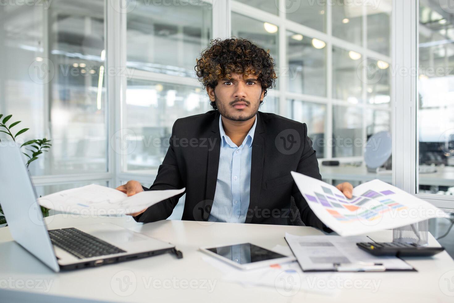 Frustrated indian male in jacket over shirt sitting by loaded table and spreading hands with documents. Young manager feeling stressed with paperwork and looking at camera with angry expression. photo
