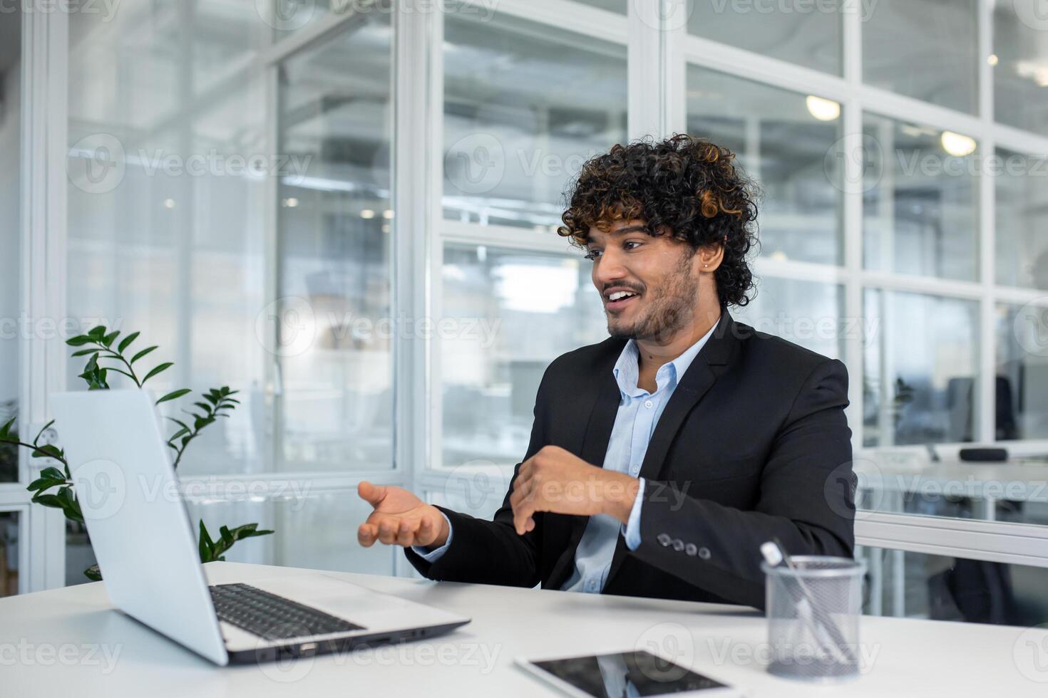 A young, curly-haired professional man in a suit engages enthusiastically in a business call, sitting in a modern office environment with natural light. photo