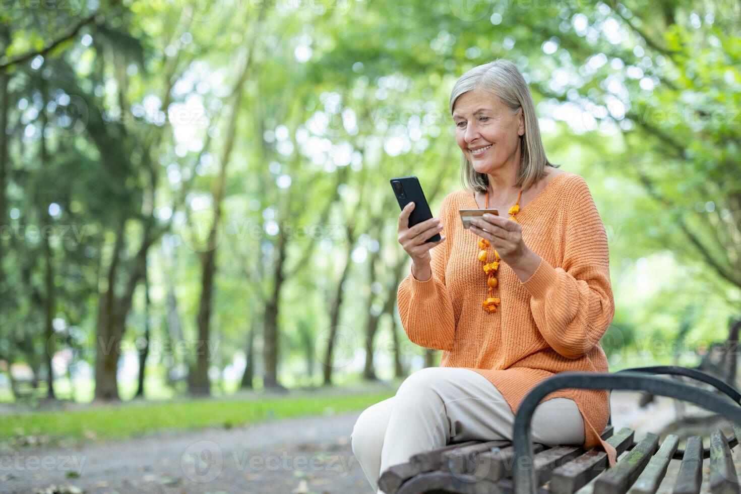 Senior mature happy woman sitting on park bench, pensioner holding bank credit card and smartphone in hands, choosing to shop online in online store, satisfaction with convenience of remote access. photo