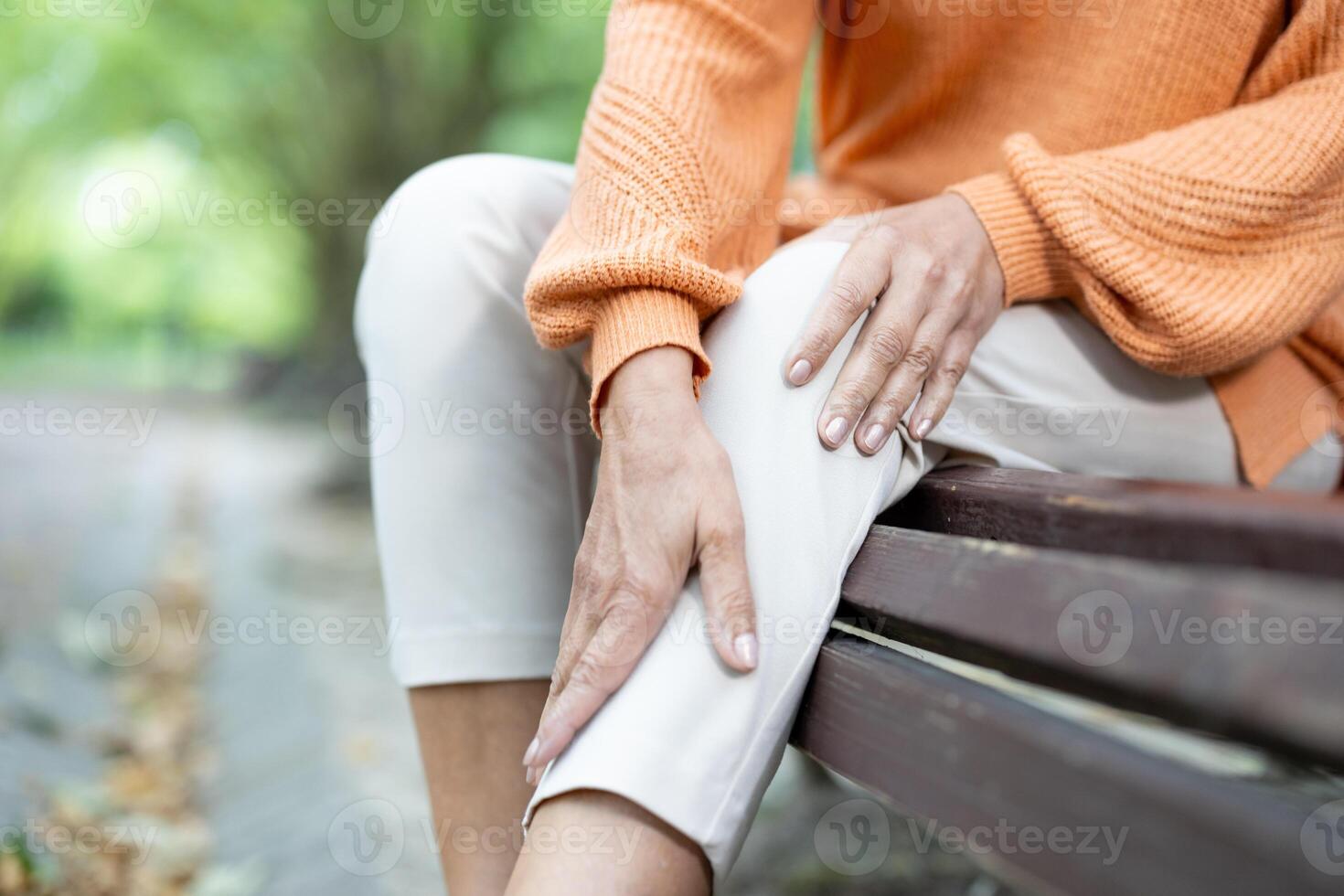 Close-up of person holding knee in discomfort, indicating knee pain. Seated on bench with sweater and light pants, scene suggests outdoor setting involving health concerns and physical discomfort. photo