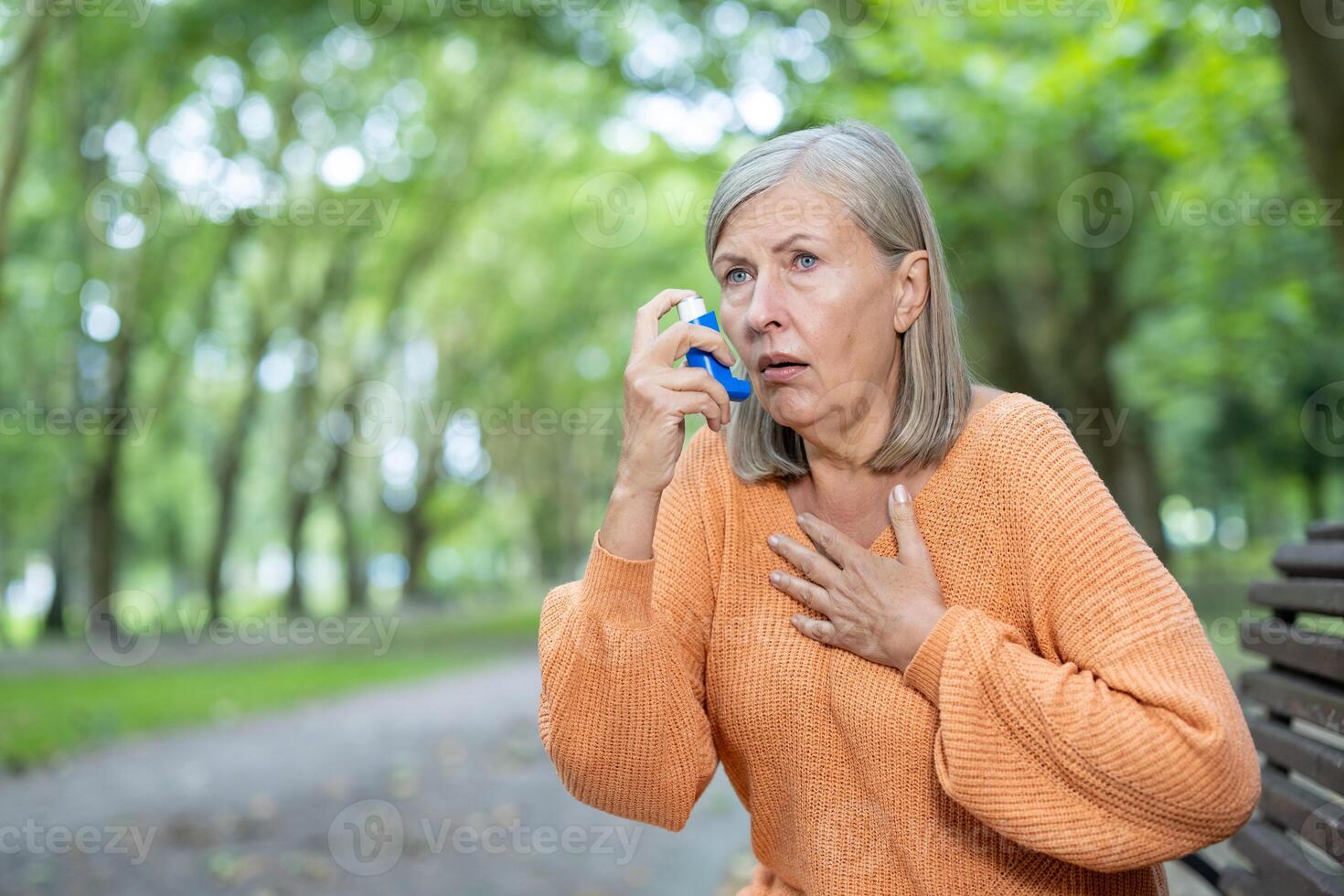 Senior woman using inhaler for asthma relief in park. Expressions of concern and calmness while managing respiratory issue. Sitting on bench, looking focused during outdoor activity. photo
