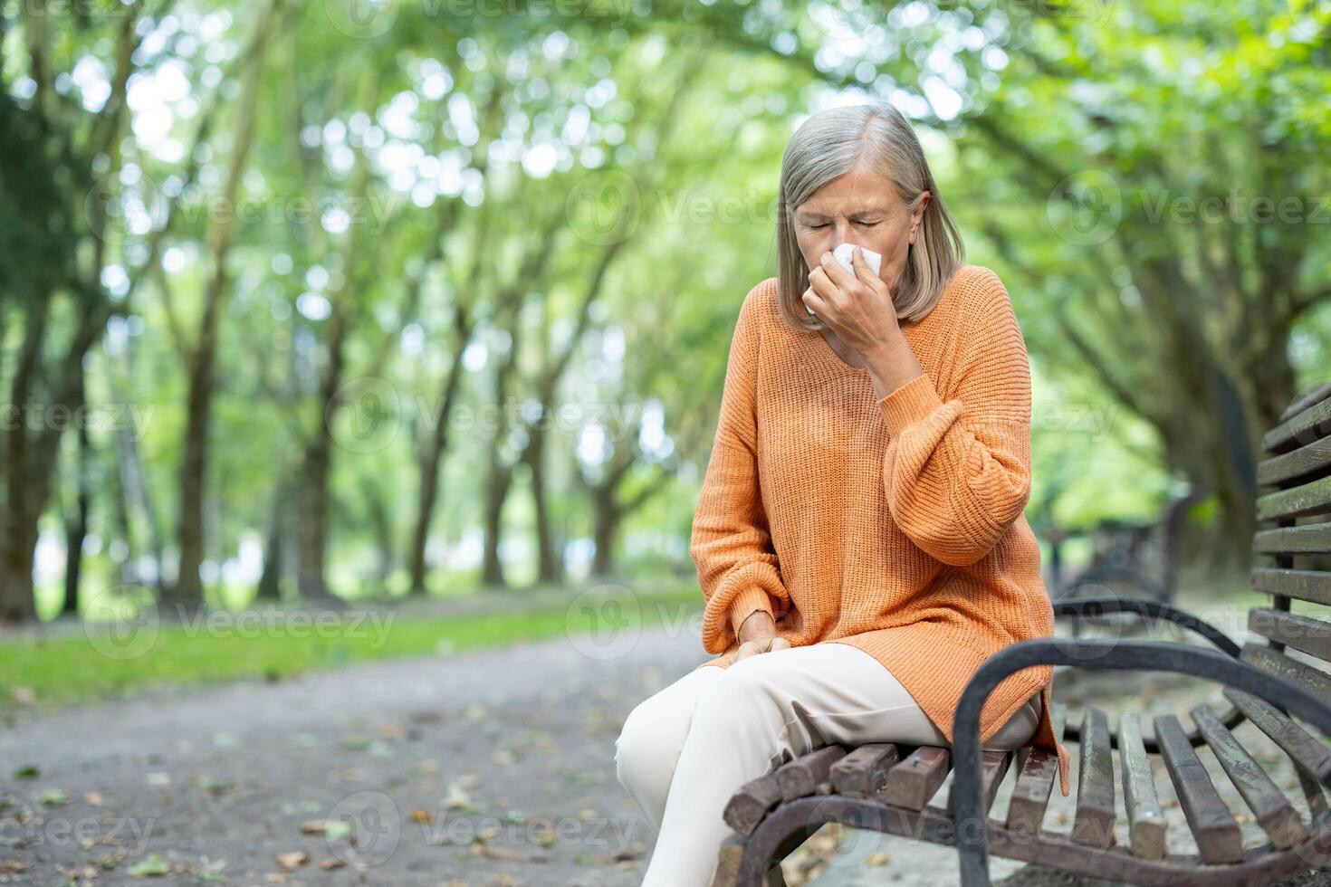 Senior woman in orange sweater sitting on park bench sneezing into tissue. Outdoor setting during autumn signifies allergy or cold symptoms, highlighting seasonal health issues and colder weather. photo