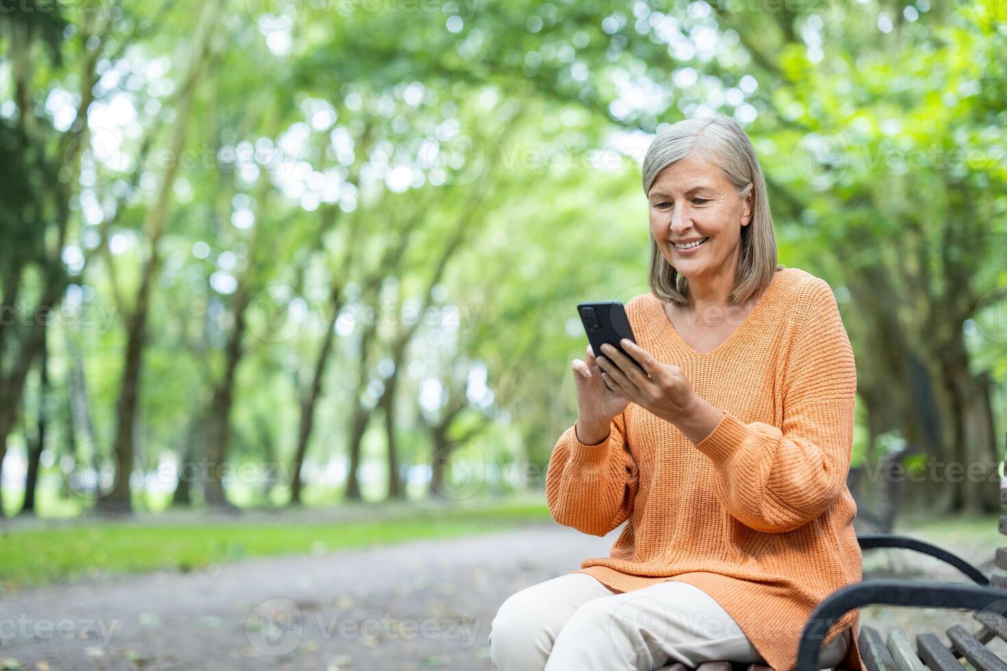 Senior woman sitting on bench in park using phone, wearing orange sweater, smiling and enjoying peaceful day outdoors, demonstrating relaxation and connection with nature and technology. photo