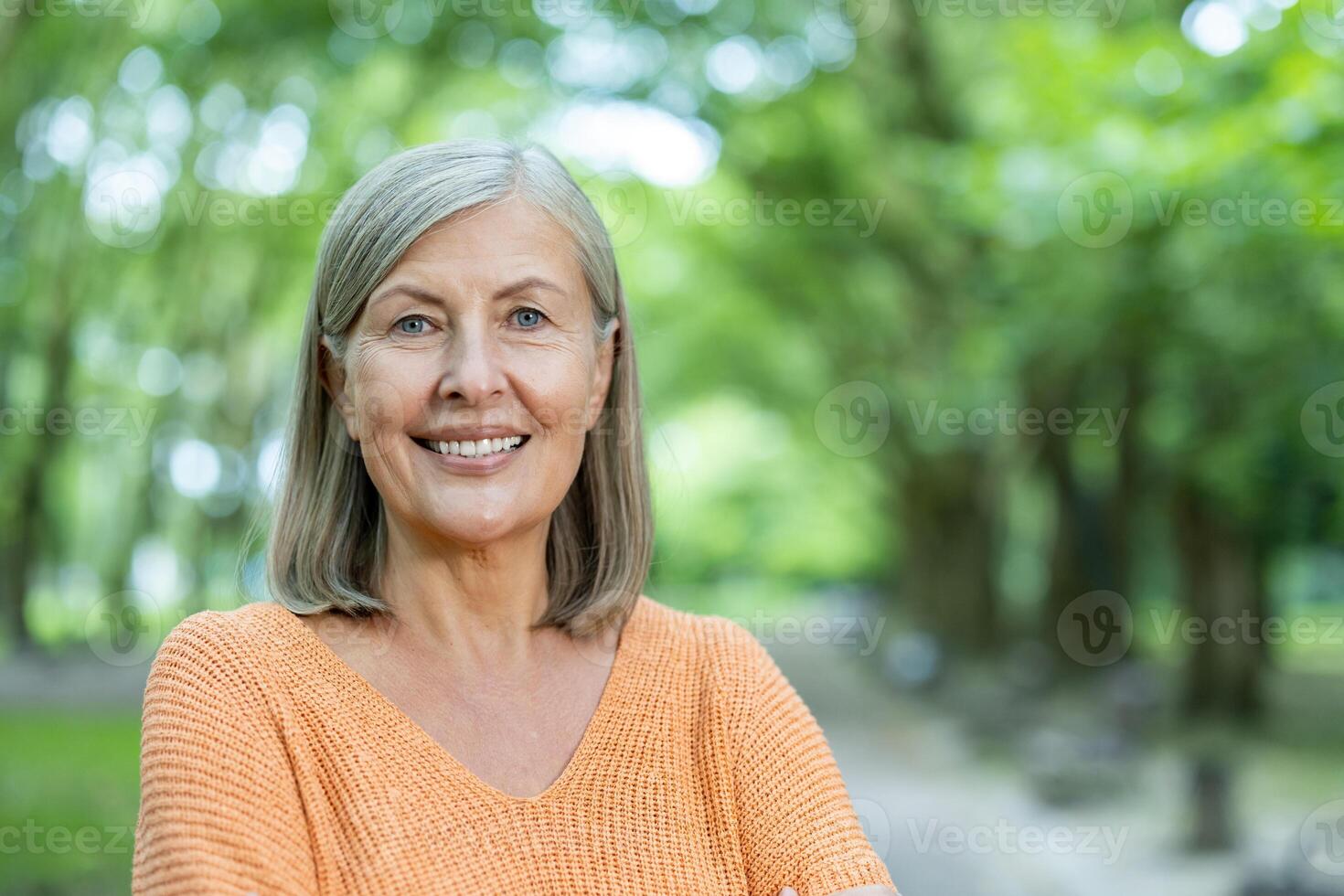 Mature woman smiles confidently standing outside . Image captures essence of positive aging and happiness. Trees and greenery create serene backdrop for this lifestyle portrait of an older adult. photo