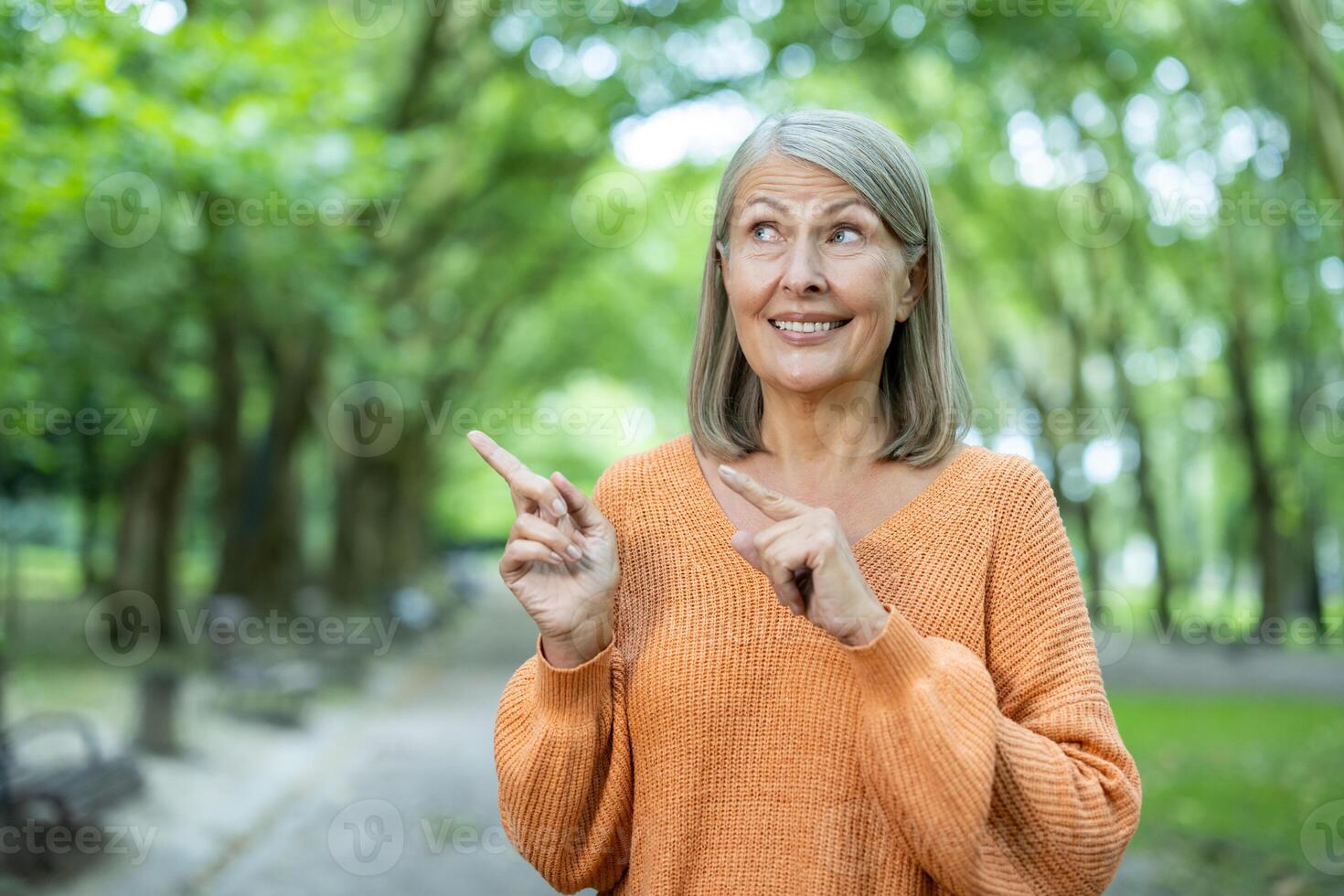 Senior woman with cheerful expression standing outdoors in park. She is wearing orange sweater and pointing fingers upwards, conveying happiness and confidence. photo