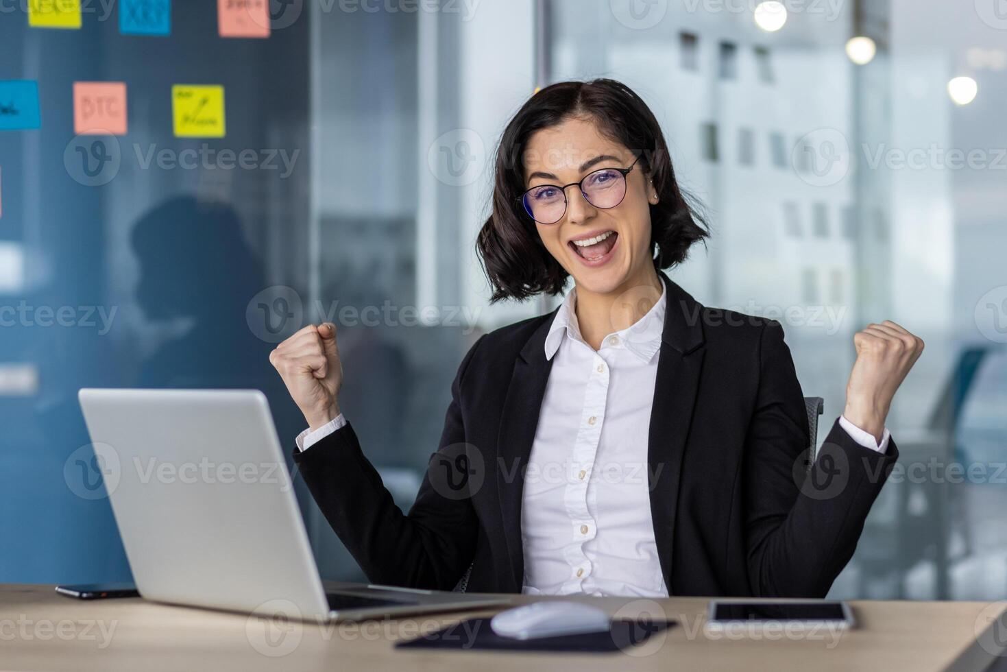 Happy businesswoman in glasses celebrating project success at office desk with laptop and phone. Expressing joy and achievement in professional environment photo