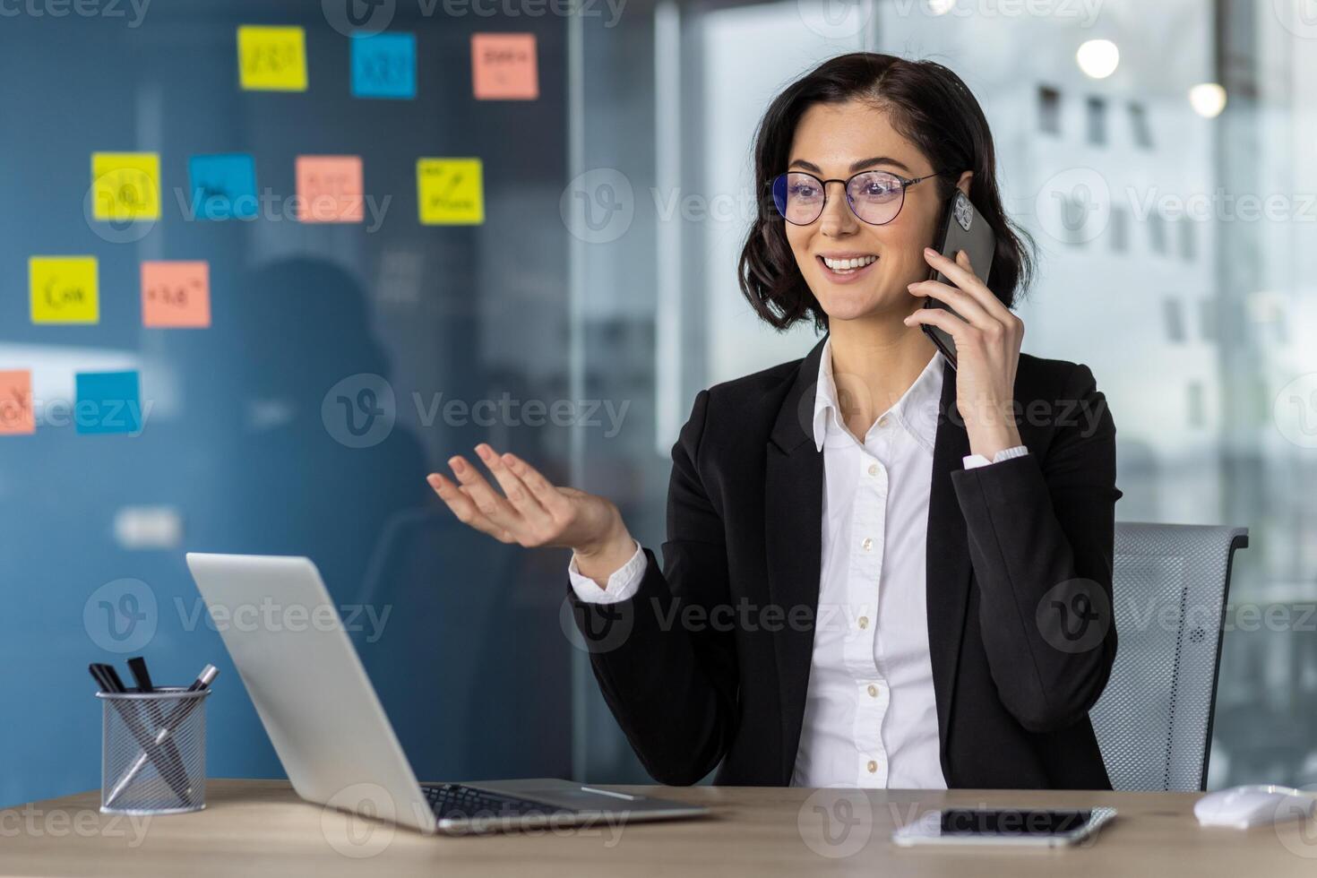 Confident businesswoman in glasses having phone call in modern office setting. Analyzing charts on laptop, smiling while discussing business strategies. Productivity and professionalism concept photo