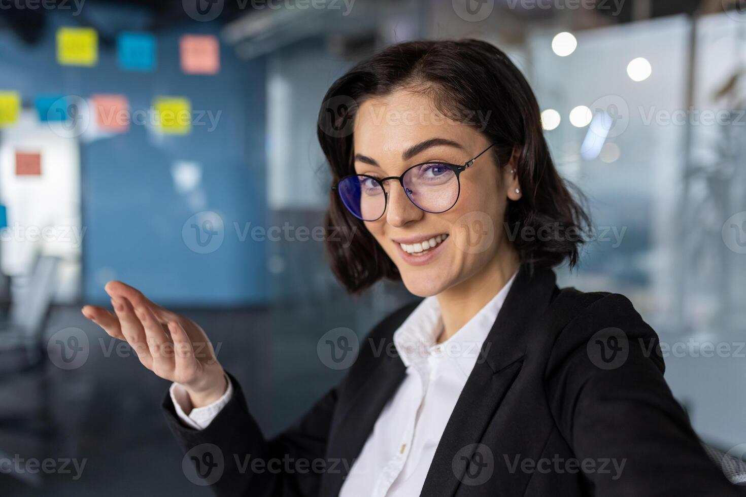 Businesswoman boss using phone for call, smiling at camera, gesturing confidently. Professional office setting with colorful notes background. Ideal for concepts of communication, technology. photo