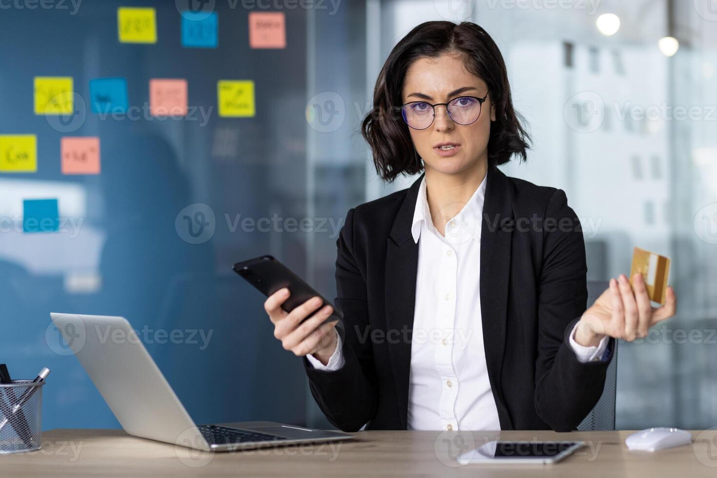 Businesswoman looking at phone and credit card with concern, considering online transaction. Office setting with laptop, highlighting concept of finance, security, and technology. photo