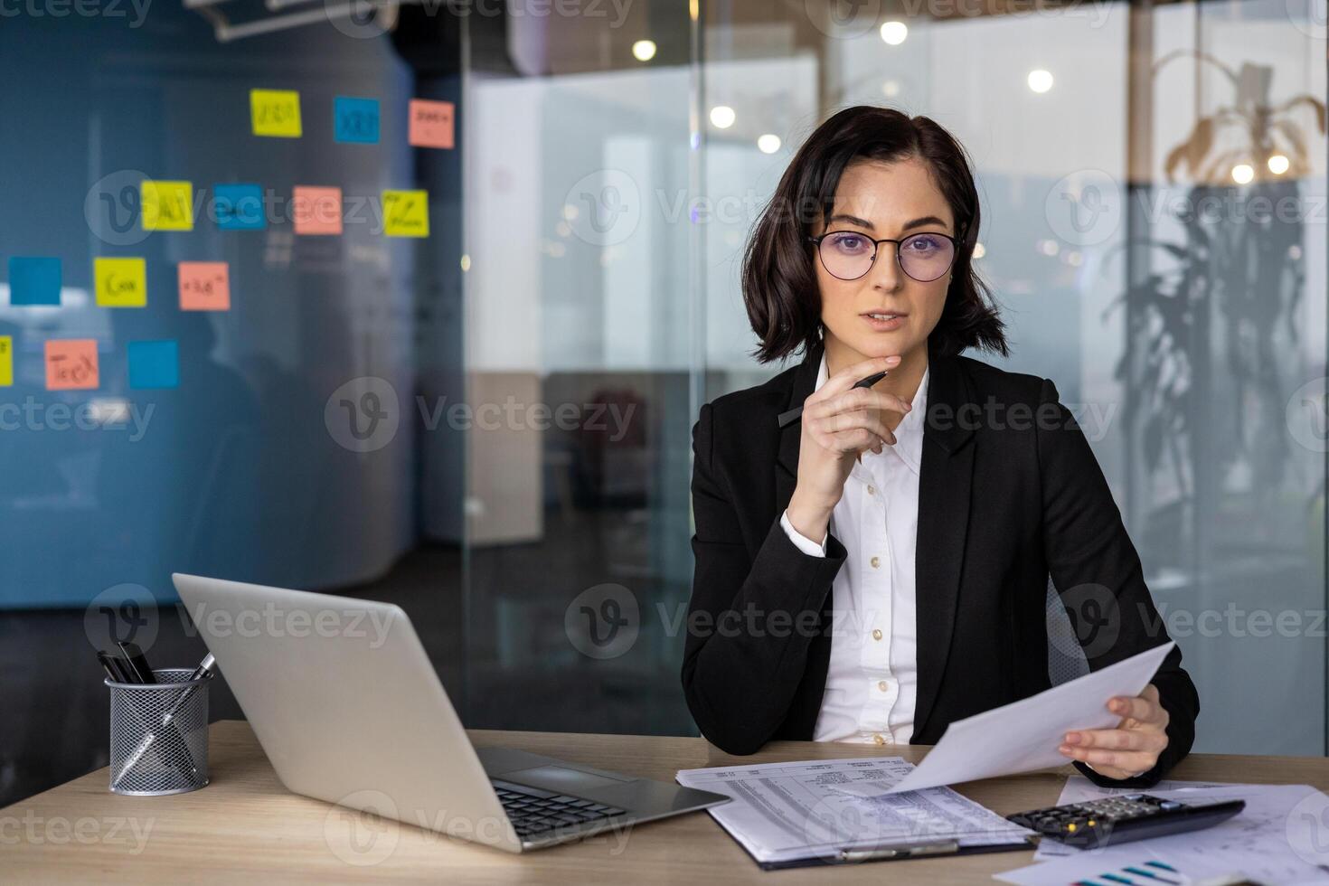Businesswoman in smart attire holds documents at desk while engaging in strategic planning. Modern office setting, with colorful notes indicating brainstorming. Use of digital tools. photo