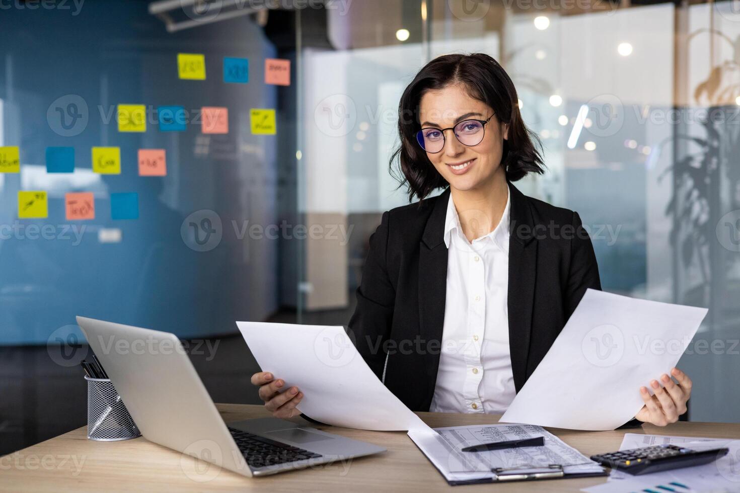 Businesswoman in office reviewing documents, demonstrating confidence and professionalism. She works at desk with laptop, showcasing leadership in workplace. Ideal for business or office themes. photo
