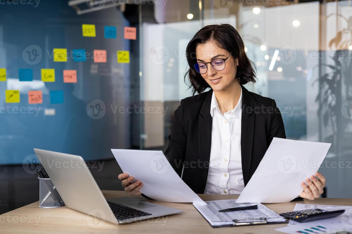 Professional businesswoman reviewing documents at desk with laptop in modern office space. Reflects focus, productivity, and confidence in corporate setting. Concept of work and strategy. photo