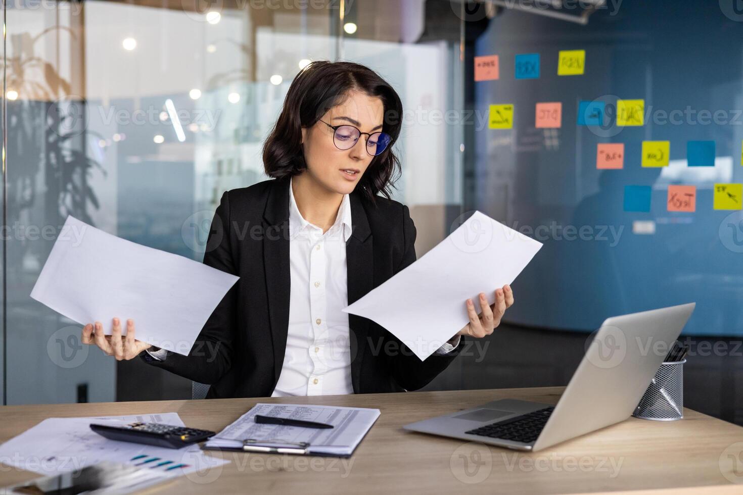 Young businesswoman in office holding papers, concentrating on data analysis, and multitasking. Wearing glasses, managing tasks at desk with laptop and sticky notes promoting productivity. photo