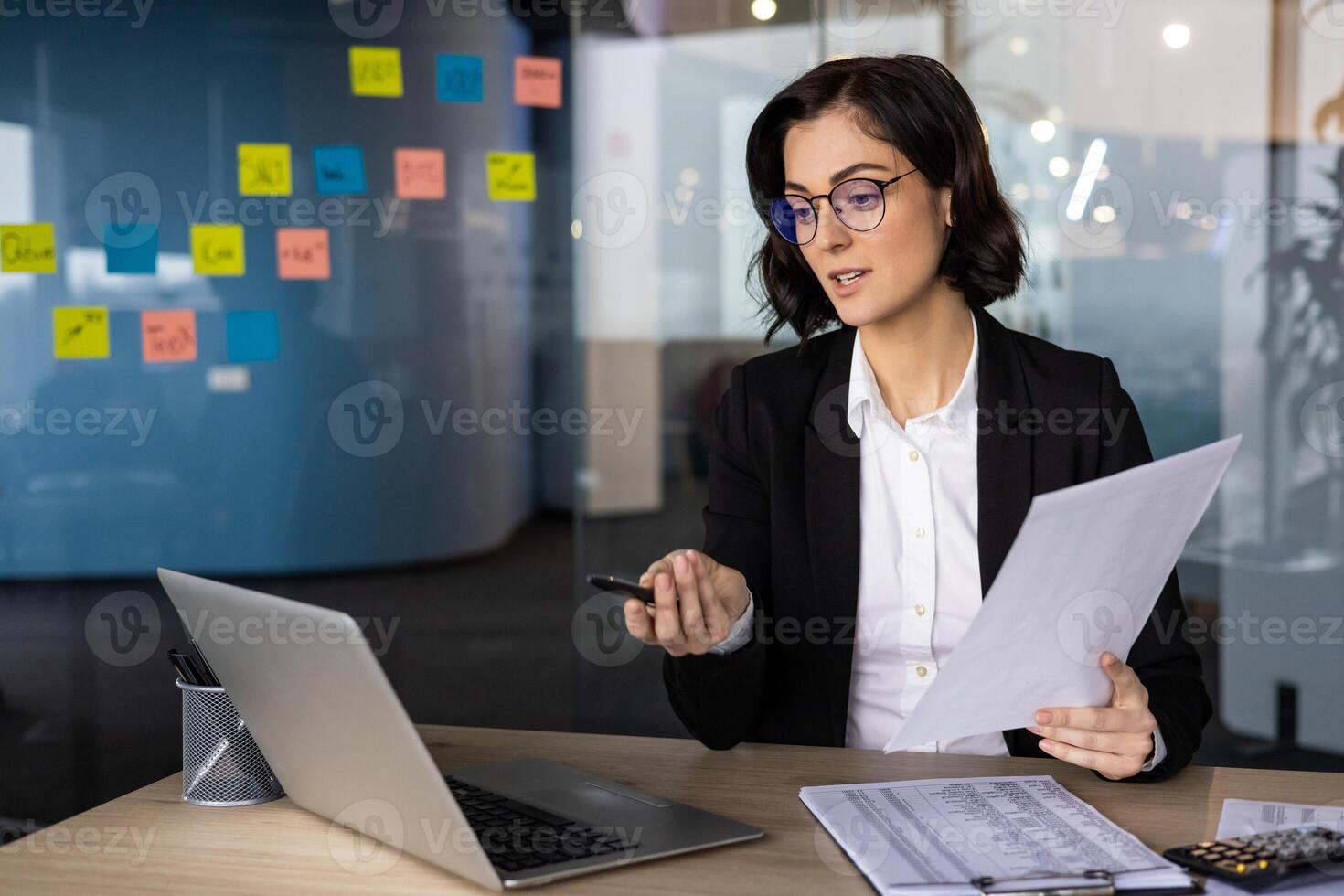 Businesswoman holding documents engages in meeting, pointing at laptop. Discusses strategies in modern office with colorful sticky notes and professional attire. Focus on collaboration, leadership. photo