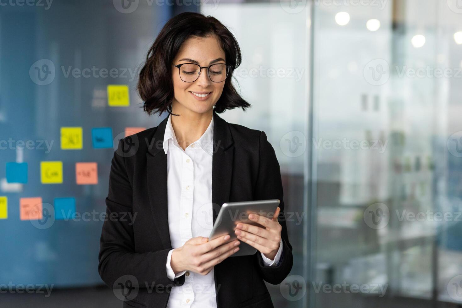 Businesswoman in office using tablet, dressed in professional attire. Confident expression shows engagement with digital tasks. Glasses add to professional appearance, promoting focus and innovation. photo