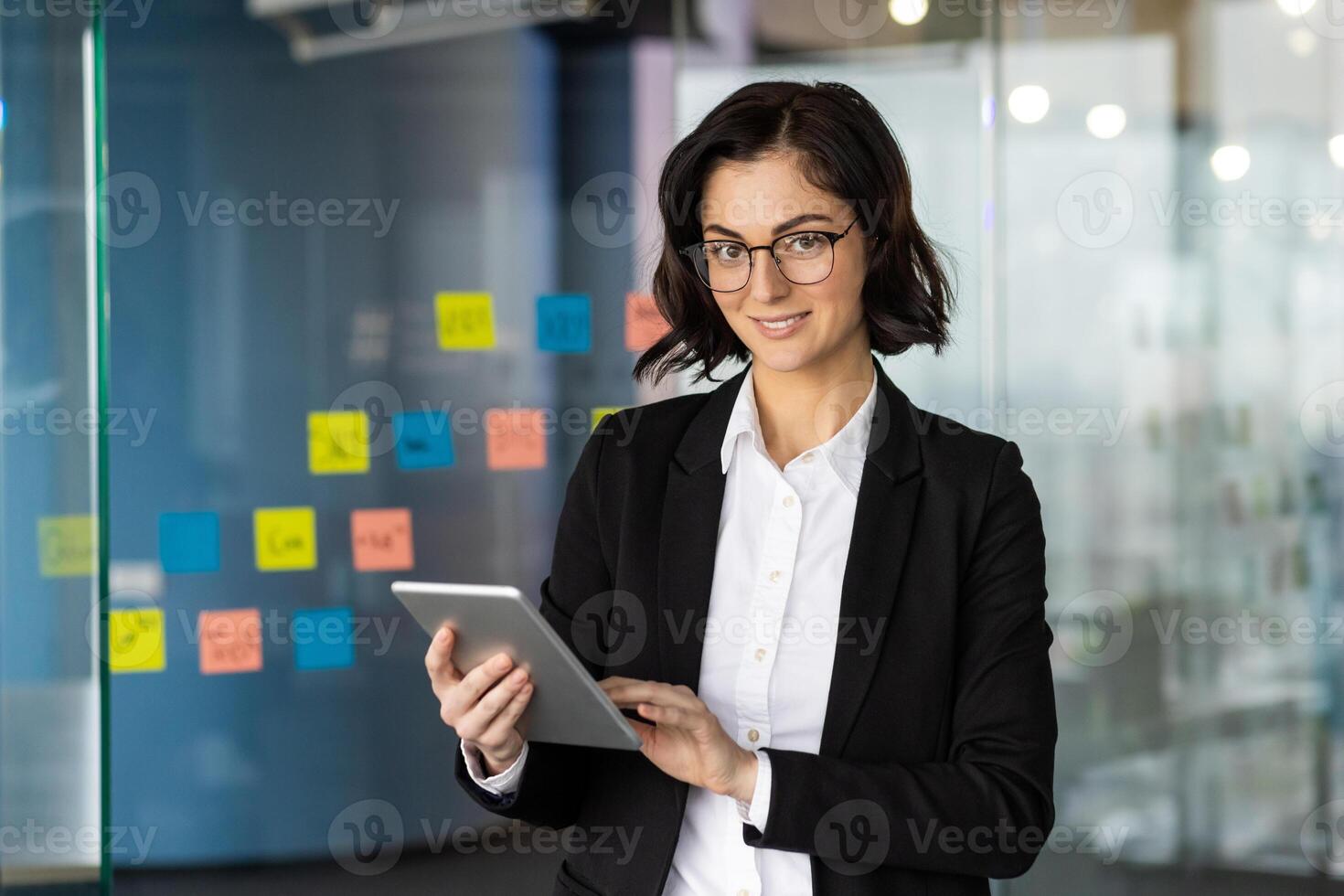Confident businesswoman using tablet in modern office setting, engaging with tasks efficiently. Brightly colored sticky notes in background highlight teamwork and innovative ideas. photo