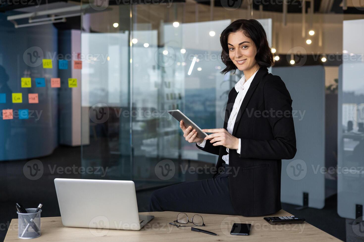 Confident businesswoman in office using tablet, smiling at camera. Surrounded by work essentials like laptop, phone, and notes on glass wall. Concept of professionalism, technology, success. photo