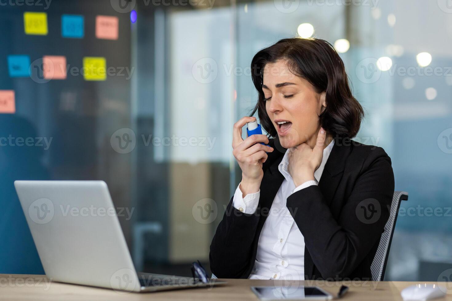 Businesswoman in office uses an inhaler for asthma relief while working at laptop. Concept of managing health at work. She is dressed in formal attire. Health and business combine. photo