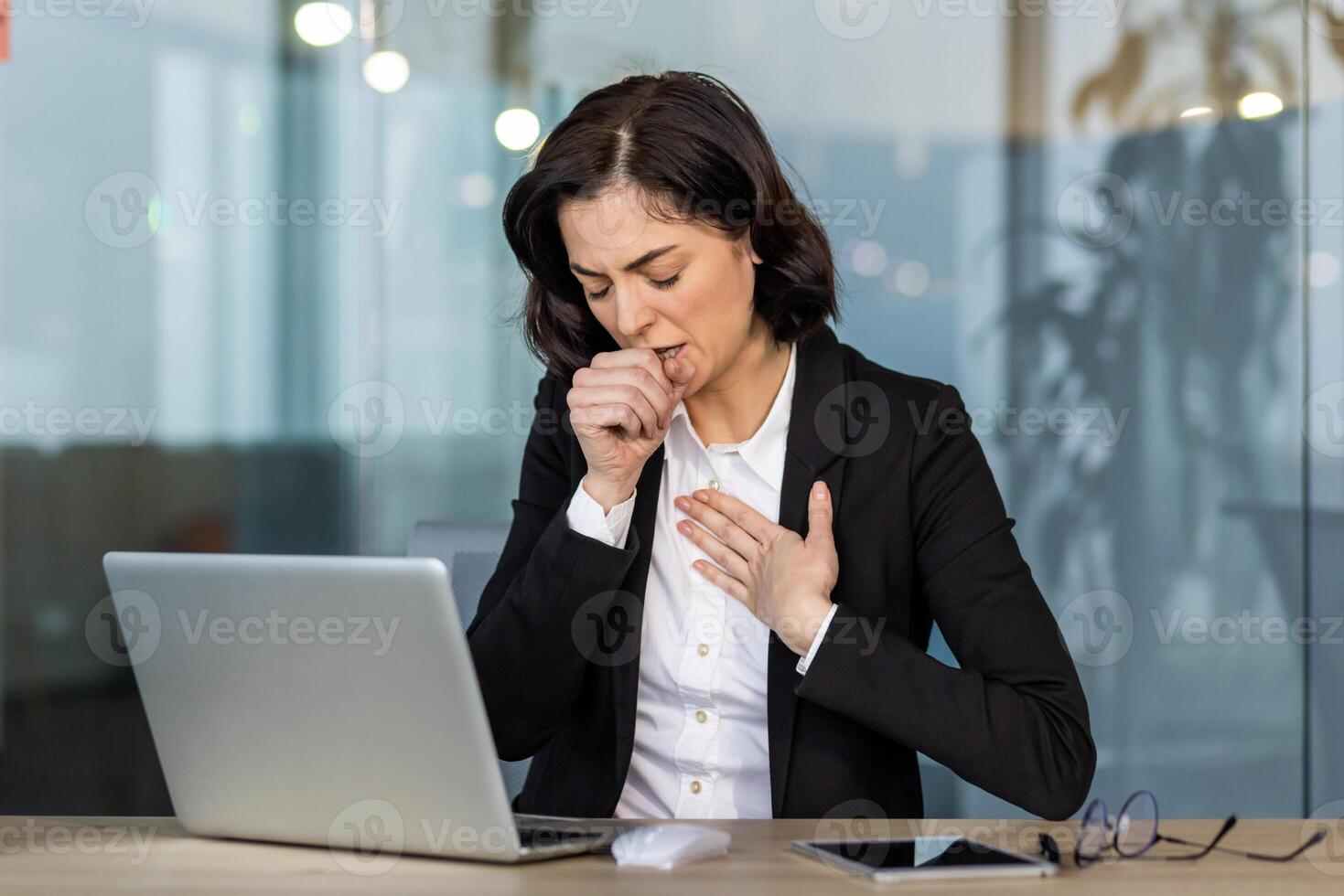 Young businesswoman wearing suit coughing while working on laptop in office setting. Health issues or discomfort during worktime illustrating common workplace challenges. photo