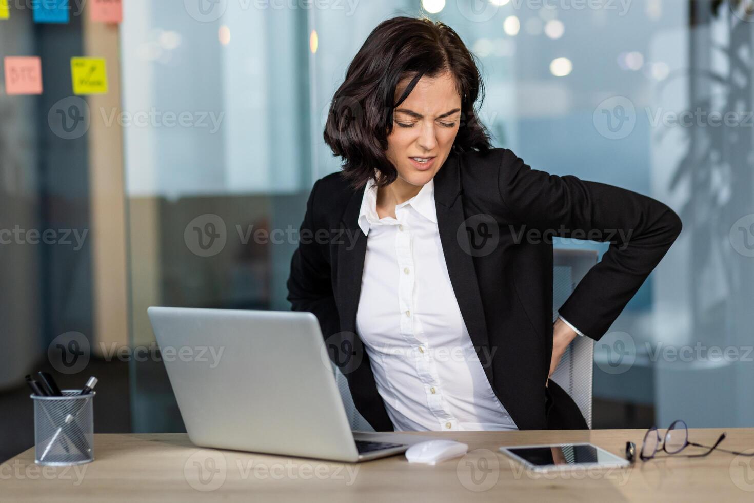Businesswoman in office showing signs of back pain while sitting at desk using laptop and phone. Indicates discomfort, poor posture, need for ergonomic solutions in workplace. photo