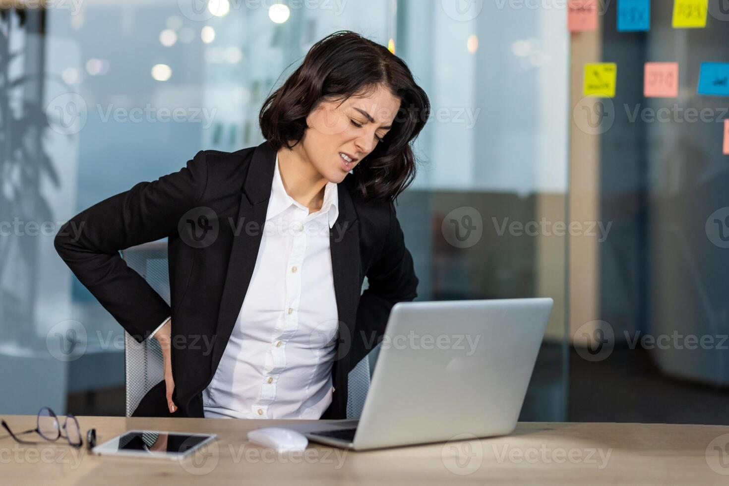 Young businesswoman in office experiencing discomfort from back pain. She is sitting at desk, with laptop and phone, displaying tension and discomfort during work hours. photo