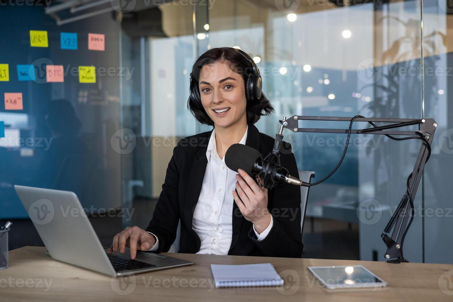 Confident podcast host in office setting, engaging with microphone. Wearing headphones, using laptop, smartphone nearby. notes display creative workspace environment, modern communication photo