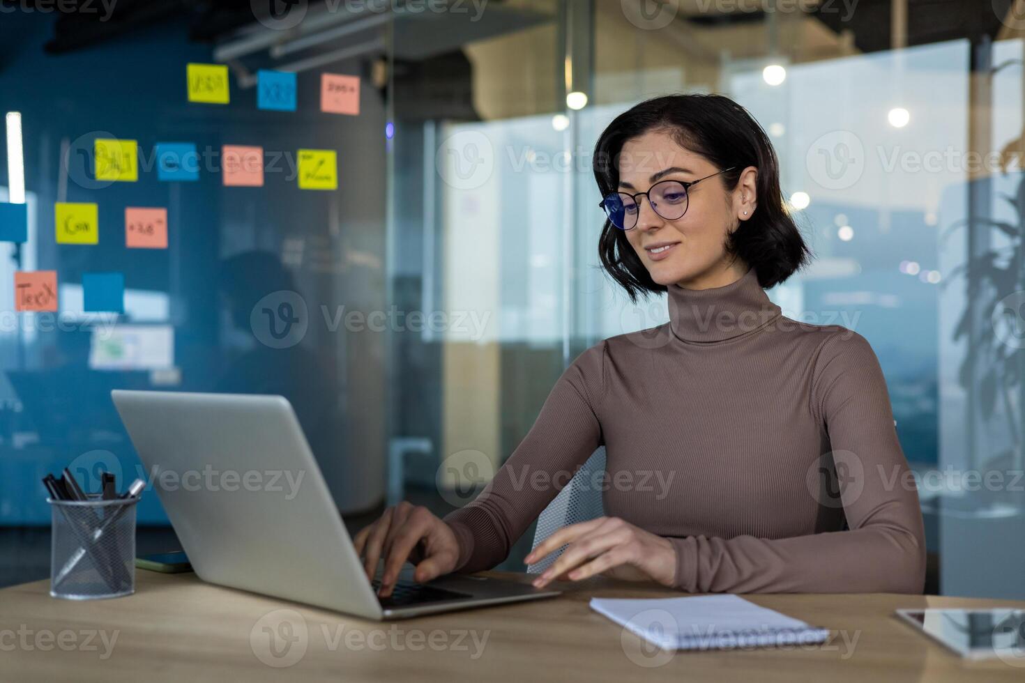 Woman working confidently on laptop in modern office setting. Focused professional in glasses using technology for business productivity. photo