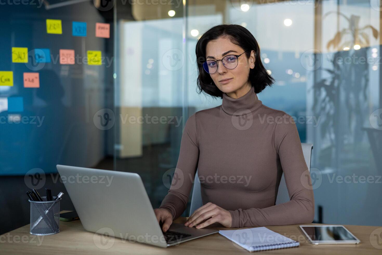 Professional businesswoman seated in modern office setting with laptop and notepad. Woman wears glasses, exuding confidence and readiness for work. Ideal for themes of business, professionalism photo