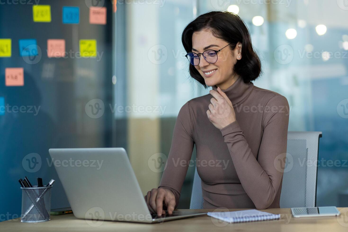 Businesswoman wearing glasses using laptop in office setting. Confidently working on tasks, showing focus and determination. Modern workspace features desk with notebook photo