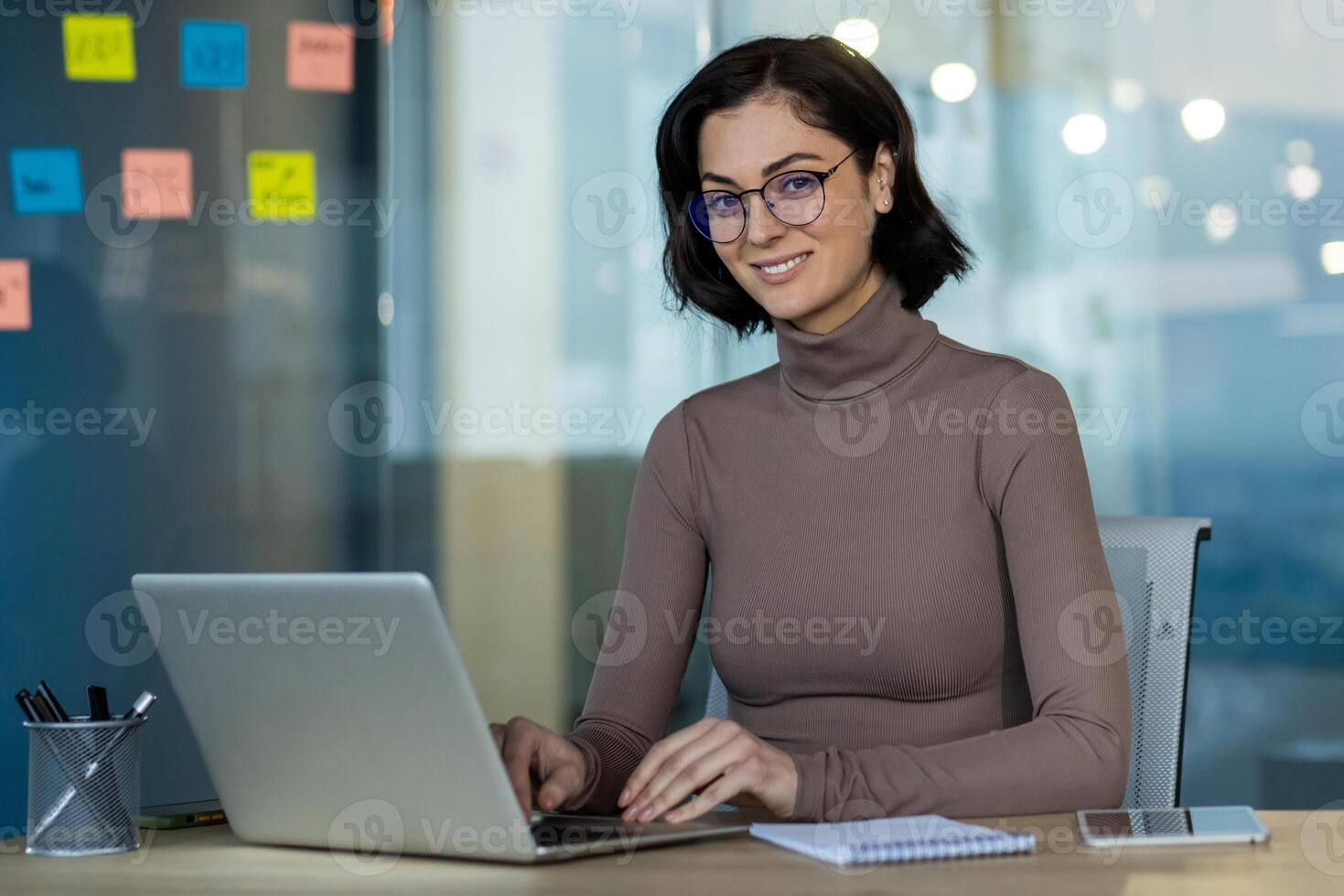Businesswoman using laptop in office demonstrates confidence and productivity. Woman with glasses sits at desk with notepad and phone, displaying professional work environment. photo