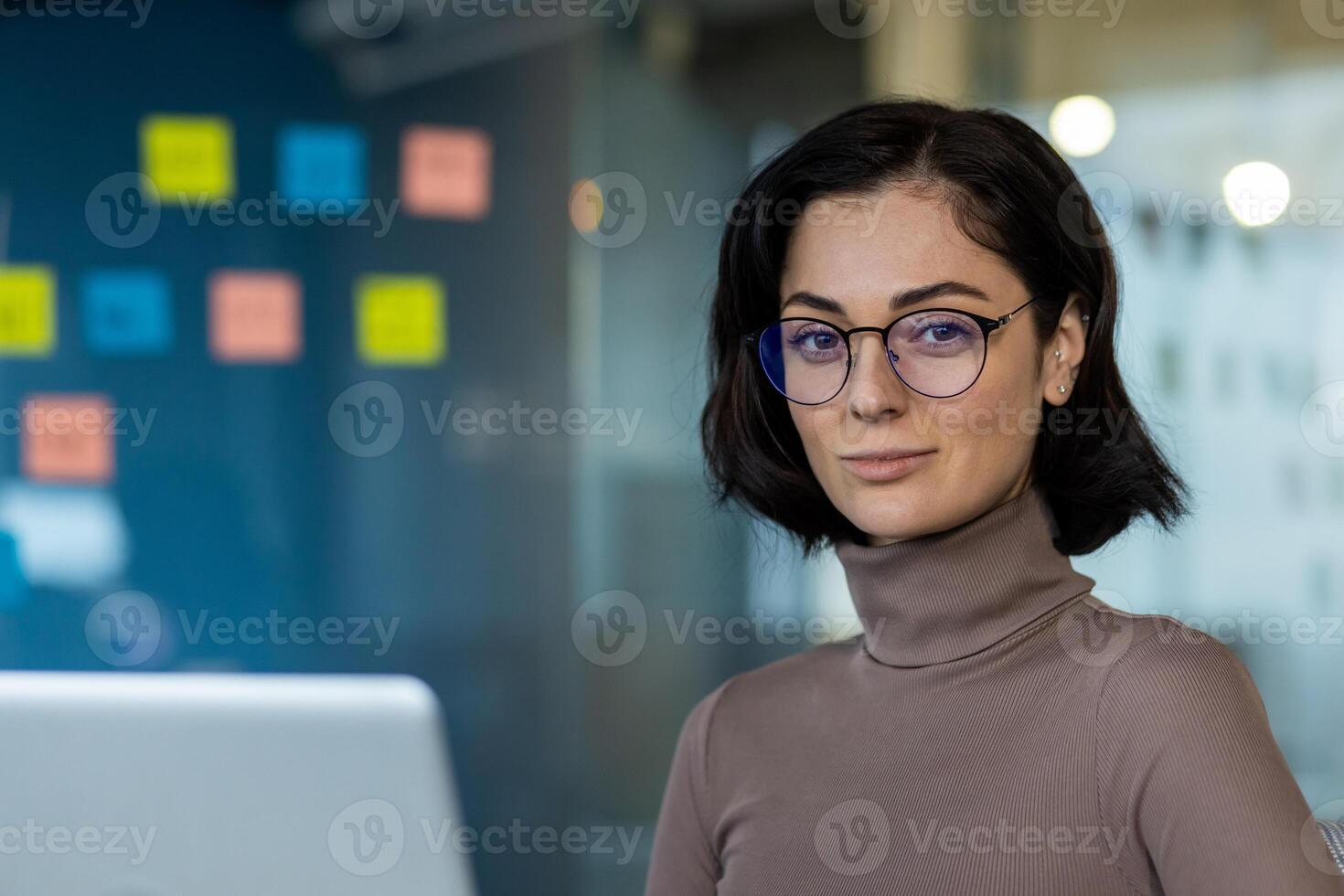 Confident woman with eyeglasses and casual attire in modern office setting. Focused professional working on laptop . Represents business, leadership, creativity, and efficiency. photo