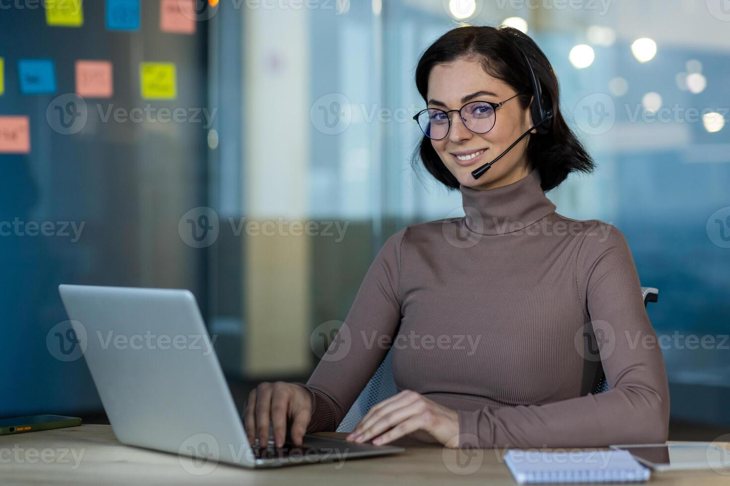 Smiling woman with glasses and headset uses laptop in office setting. Engaged in online meeting or support call. Modern technology enhances communication and productivity in business environment. photo
