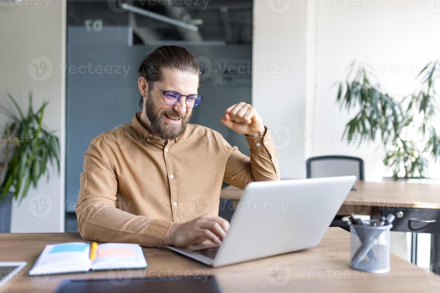 A young man with glasses and a beard expressing excitement working on a laptop, showing a celebratory fist. Perfect for depicting success, achievement, and positivity in a professional workspace. photo