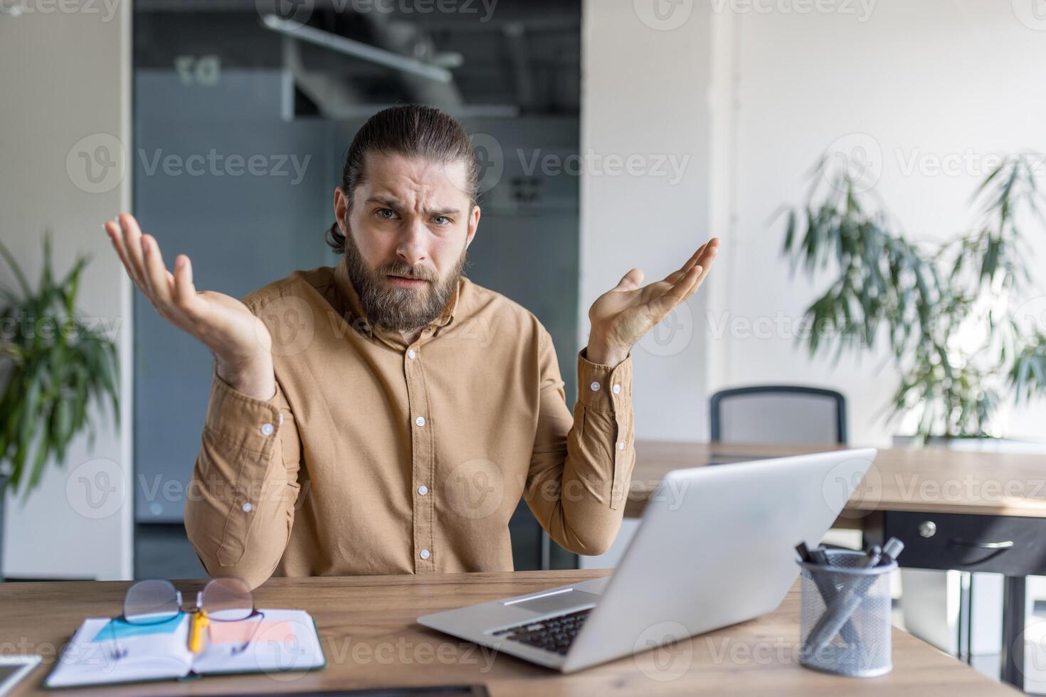 A man with a puzzled expression sits at a desk using a laptop, gesturing with raised arms, conveying confusion or hesitation while working in an office or professional setting. photo