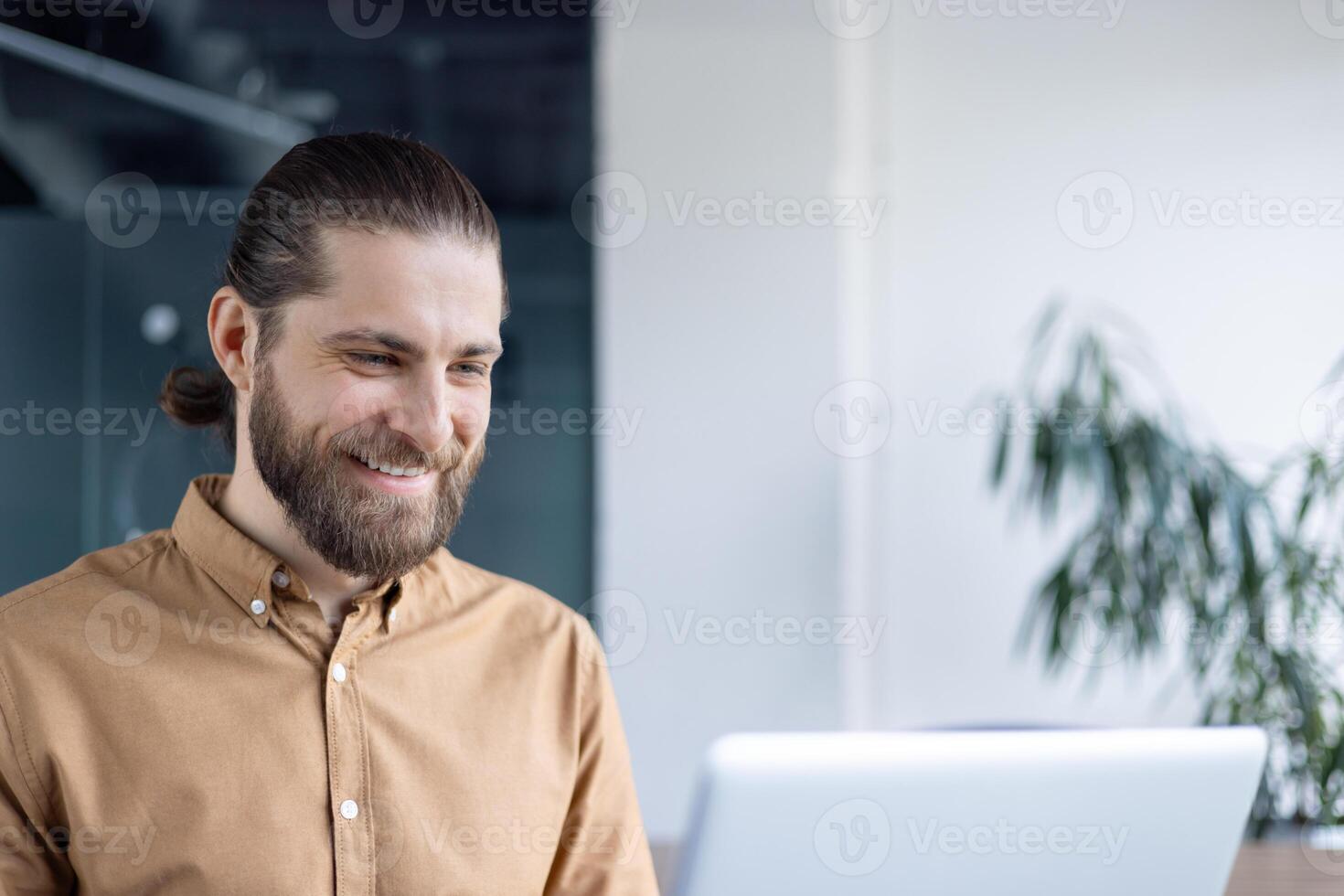 A cheerful man with a beard uses his laptop while seated in a modern office setting. The image conveys a positive mood, focusing on productivity and workplace happiness. photo
