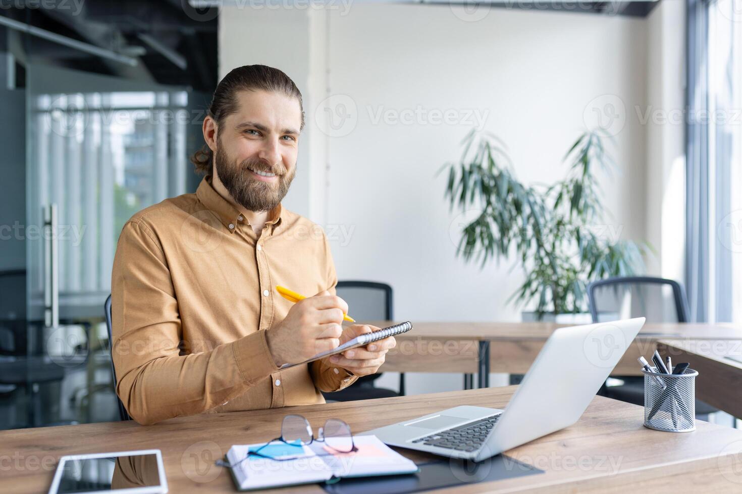 Confident professional taking notes while working on a laptop at a modern office desk, representing productivity and creativity in a workspace. photo