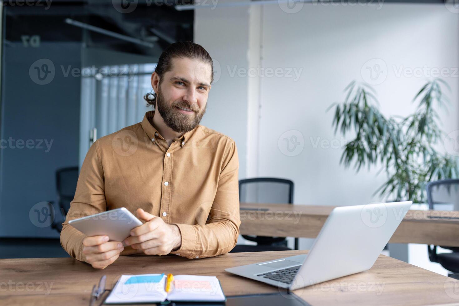 Confident young man using a digital tablet at work with a laptop and notebook, exuding professionalism and focus in a modern office environment. photo
