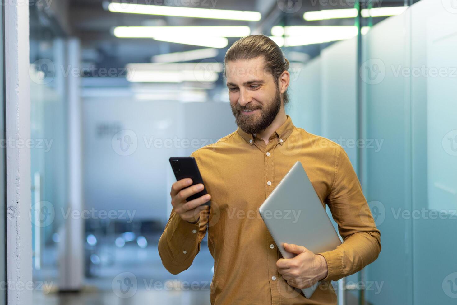 A professional man stands in a modern office setting, smiling at his smartphone while holding a laptop. The image conveys productivity and a positive workplace atmosphere. photo
